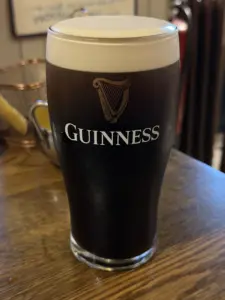 A Guinness pint glass with a creamy head sits on a wooden table, showcasing the iconic logo and harp. The blurred background evokes the warmth of a cozy pub setting.