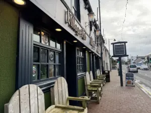 Wooden chairs are lined up outside a pub with a sign reading "Village Inn" and a Guinness pint glass displayed nearby. Another sign down the street says "Mary's Village Pub Restaurant." The quiet street has a few cars parked along the road.