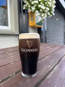 A pint of Guinness stout with a creamy head sits on a weathered wooden outdoor table, with flowers, a yellow box, and a closed metal shop shutter in the background.