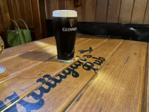 A Guinness pint glass rests on a wooden pub table with “Arllugair’s Pub” in black script. The background features wooden paneling and a green bag on a bench.