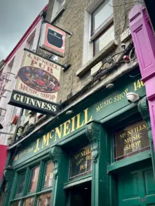 Street view of J. McNeill Music Shop and Pub in Dublin, featuring green storefront, gold lettering, a hanging pub sign, Guinness sign, and neighboring brick buildings.