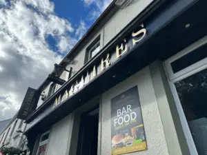 Low-angle view of the exterior of Maguires pub, with a black and gold sign and a poster advertising bar food, including a burger and fries, on a white wall. The sky is partly cloudy above.