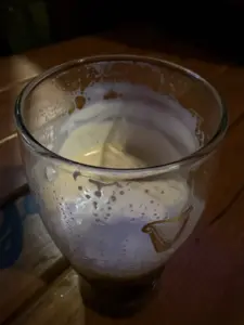 A nearly empty Guinness pint glass with remnants of foam sits on a wooden table, showing lacing around the inside and a faint harp logo, evoking the atmosphere of a cozy pub.