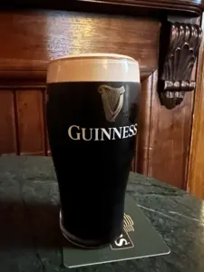 A pint of Guinness stout with a creamy head sits on a green coaster on a dark table in a wood-paneled pub.