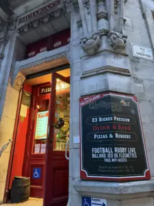 Entrance of Mulligan's Pub on a stone street corner, with a blackboard sign featuring a guinness pint glass, food, live sports, and free WiFi. The open pub door glows warmly inside. French street signs are visible.