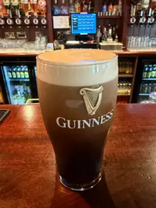 A Guinness pint glass with a thick, creamy head sits on a wooden bar counter; behind it, shelves hold bottles, glasses, and a register in a cozy pub setting.