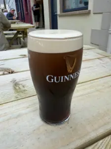 A Guinness pint glass with a creamy head sits on a weathered wooden outdoor table, with buildings and people blurred in the background, evoking the lively atmosphere of a pub.