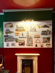 A large painting of various buildings and landmarks, including a Guinness pint glass pub, hangs on a dark green and red wall above a cream-colored fireplace in a cozy room with red ceiling and ornate chandelier.