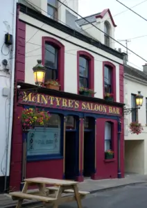 McIntyre's Saloon Bar is a traditional pub with cream and burgundy trim, hanging flower baskets, old-fashioned lamps, and a wooden picnic table outside on a quiet street—perfect for relaxing with a Guinness pint glass in hand.
