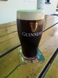 A Guinness pint glass with a creamy head sits on a coaster at a pub’s wooden outdoor table, sunlight casting reflections across the surface.