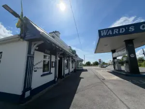 A small white building labeled "The Lounge Entrance" stands beside Ward's Oil gas station under a bright, sunny sky, hinting at a welcoming pub atmosphere with a Guinness pint glass sign visible by the door.