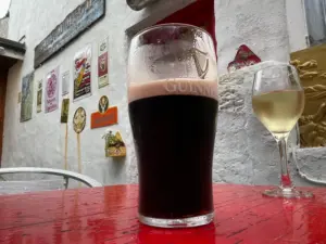 A Guinness pint glass and a glass of white wine sit on a red table outside a pub, with a white wall decorated with various signs and plaques in the background.