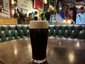 A Guinness pint glass with a creamy head sits on a wooden table in a cozy pub, featuring green tufted seating and colorful decorations in the background.