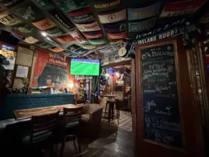 A cozy pub with wooden tables and chairs, rugby scarves covering the ceiling, a chalkboard menu at the entrance, and a Guinness pint glass on the table in a dimly lit, inviting atmosphere.
