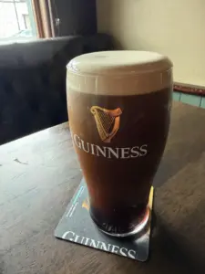 A freshly poured Guinness pint glass with a thick, creamy head sits on a coaster atop a wooden table in a dimly lit pub.