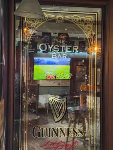 A glass door with ornate gold designs reads "The Oyster Bar" and "Guinness 1759." Through the door, a TV shows a sports game inside a cozy pub with wooden tables, chairs, and a Guinness pint glass on the counter.