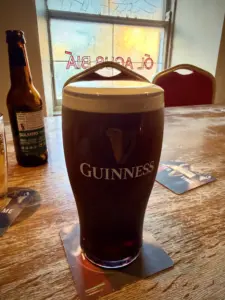 A Guinness pint glass with a creamy head sits on a wooden table near a window in a pub, accompanied by a bottle of Bulmers cider and red chairs in the background.