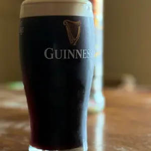 A Guinness pint glass with a creamy head sits on a wooden surface in a pub setting, with the iconic Guinness logo and harp emblem visible on the glass against a blurred background.