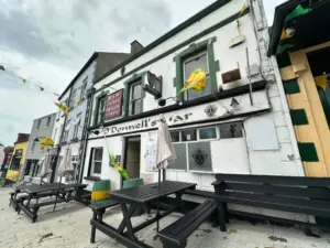 Street view of O'Donnell's Bar with green and yellow flags, outdoor wooden benches, a whitewashed exterior, and a classic Guinness pint glass logo on display, located in a row of buildings under a cloudy sky.