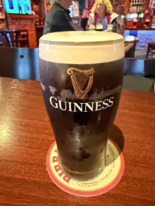A Guinness pint glass with a creamy head sits on a branded coaster atop a wooden table in a pub, with people and bar decor visible in the background.