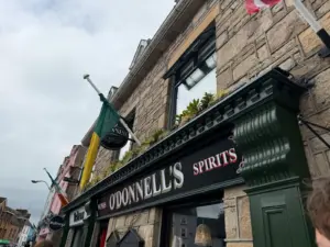 A close-up of O'Donnell's Spirits shop, a stone building with flags and a Guinness pint glass on the sign, under a cloudy sky. Green trim decorates the shopfront; other colorful buildings line the street.