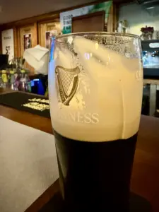 A pint of Guinness with a creamy head sits on a bar counter, with bar equipment and drink taps visible in the softly lit background.