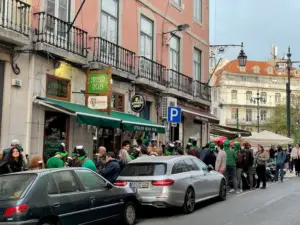 A crowd of people, many wearing green hats and shirts, gather outside an Irish pub on a city street lined with parked cars and historic buildings.