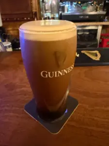 A pint of Guinness with a creamy head sits on a coaster on a wooden bar, with bar equipment and a harp logo visible in the background.