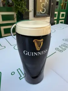 A pint of Guinness stout sits on an outdoor table, featuring a creamy head and the iconic harp logo on the glass. The background shows part of a pub with green doors and windows.