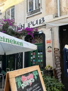 A charming pub with a green door and sign reading "PUB" sits beneath vibrant purple flowers. A Heineken umbrella and chalkboard menu stand outside, with clothing and accessories displayed to the right.