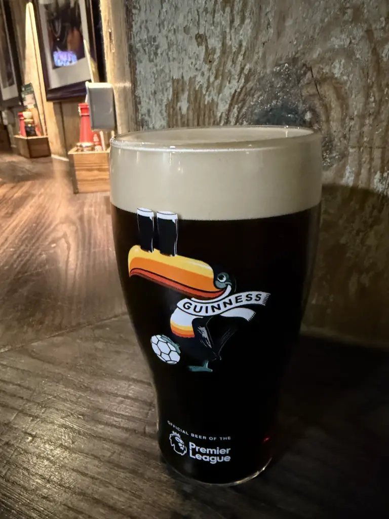A guinness pint glass with a creamy head sits on a wooden table in a pub. The glass features a toucan, a soccer ball, and the Premier League logo, with framed photos and rustic decor visible in the background.