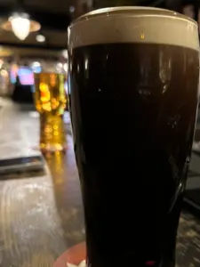 A close-up of a dark beer in a pint glass sits on a bar counter, with a blurred background showing another glass of light beer and dim bar lighting.