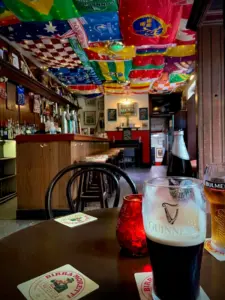 A cozy pub interior with international flags hanging from the ceiling, a bar on the left, and a table in the foreground featuring a Guinness pint glass, a cider, coasters, and a red candle.