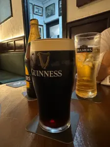 A Guinness pint glass sits on a pub table next to a Bulmers Irish cider glass and bottle, with cozy pub decor and framed photos on the walls in the background.