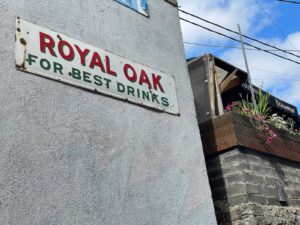 A weathered sign on a textured gray wall reads "ROYAL OAK FOR BEST," evoking a classic pub vibe. Nearby, a flower box with pink flowers sits on a stone wall under a sunny, partly cloudy sky, hinting at the charm of a Guinness pint glass waiting inside.