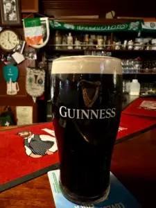 A Guinness pint glass with a creamy head sits on a bar counter in a cozy pub decorated with Irish flags, scarves, and memorabilia in the background.