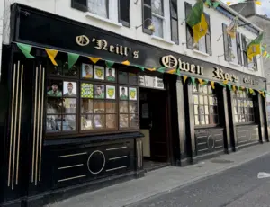 The exterior of "O’Neill’s Owen Roe’s" pub features yellow and green flags, banners, and posters with faces in the windows celebrating a sports event. A guinness pint glass design is displayed on the black and white painted walls.