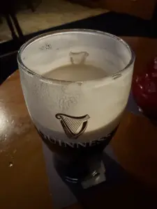 A partially finished Guinness pint glass sits on a coaster atop a wooden table in a dimly lit pub. The creamy foam leaves lacing on the glass, with the Guinness logo clearly visible.