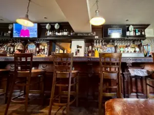 A wooden bar with high chairs, beer taps, bottles, and glasses. Two ceiling lamps hang above. A TV on the left shows a news anchor; another screen on the right displays a blank message. The bar is empty of people.