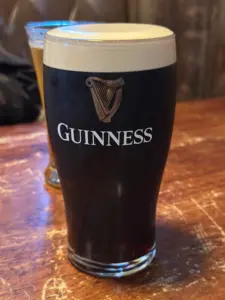 A pint glass of Guinness stout with a creamy head sits on a wooden table; another drink is blurred in the background. The Guinness logo and harp are visible on the glass.