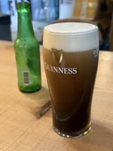 A pint glass of Guinness with a creamy head sits on a wooden table next to an empty green glass bottle. The background is blurred, suggesting an indoor setting, possibly a bar or pub.