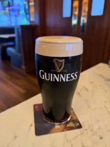 A Guinness pint glass with a creamy head sits on a bar coaster atop a marble countertop in a warmly lit pub.