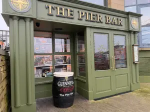 Exterior of The Pier Bar with green facade, gold lettering, glass doors, and windows displaying photos. A large black Guinness pint glass stands outside the entrance, emphasizing its iconic pub atmosphere.