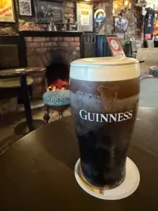 A Guinness pint glass sits on a table in a cozy pub with vintage decor, a brick fireplace with a glowing fire, and various old signs on the walls in the background.