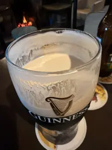 A partially finished Guinness pint glass rests on a coaster atop a table in a cozy pub, with foam clinging to the inside of the glass. In the background, there’s a warm fireplace and a bottle on display.