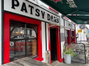 Street view of Patsy Dan’s Bar with bold black lettering, red and white exterior, arched window, and outdoor seating under green Heineken umbrellas. A Guinness pint glass is displayed beside the pub's clock and ATM on the wall.