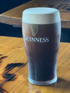 A pint of Guinness stout with a creamy head sits on a wooden table, displaying the Guinness logo and harp on the glass.
