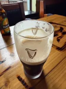A half-full pint glass of Guinness sits on a wooden pub table, with foam clinging to the inside. In the background, there is a bottle of Bulmers cider and empty chairs.