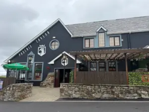 A grey building with a sign reading "Phoenix Tavern" on the wall, a wooden pergola, stone walls, hanging plants, and outdoor seating under a green umbrella. The sky is cloudy.