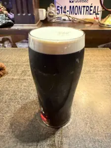 A pint of dark beer with a creamy head sits on a wooden table in a pub, with a Quebec license plate labeled "514-MONTRÉAL" visible in the background.